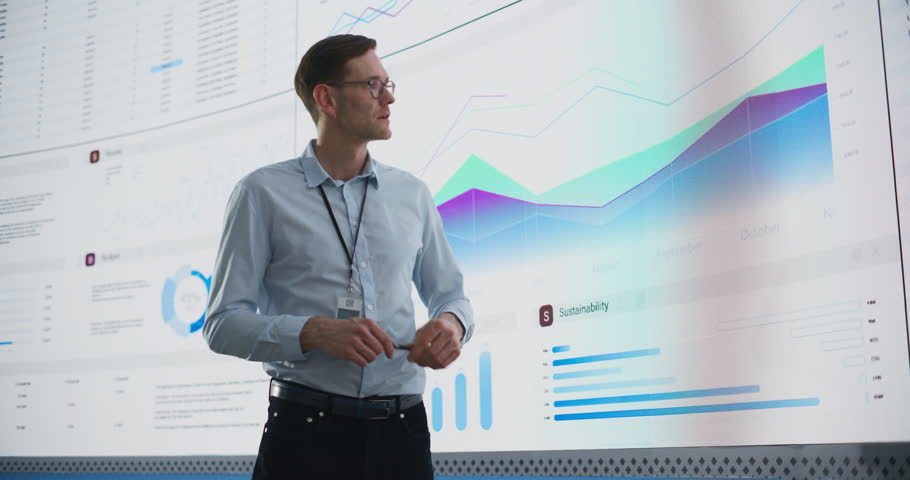 Male CEO Of Data Science Company Is Giving Presentation In Front Of Big Digital Screen With Graphs And Charts. Successful Caucasian Man Reviewing Business Accomplishments And Objectives In the Office. Royalty-Free Stock Footage #1103393029