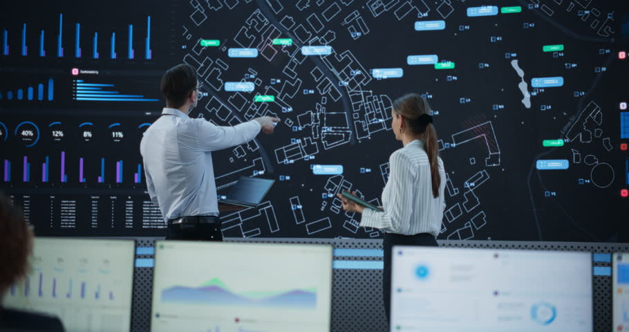 Male And Female Logistics Specialists Discussing New Routs Of Delivery In Front Of Big Digital Screen In Monitoring Office. Diverse Employees Working Behind Desktop Computers For Startup Service.