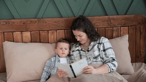 Young woman reads a book with fairy tales to her toddler son before going to bed. The child reads a book with his mother lying in bed. Happy childhood. Slow motion video