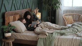 Young woman reads a book with fairy tales to her toddler son before going to bed. Child and his mom in cute pajamas reads a book lying in bed. Happy childhood. Slow motion video