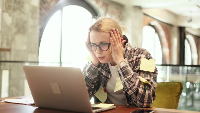Nervous stressed young blond woman freelancer with stickers typing quickly on laptop computer and looking at paper documents asking to hurry up scream and warning about deadline at coworking workplace