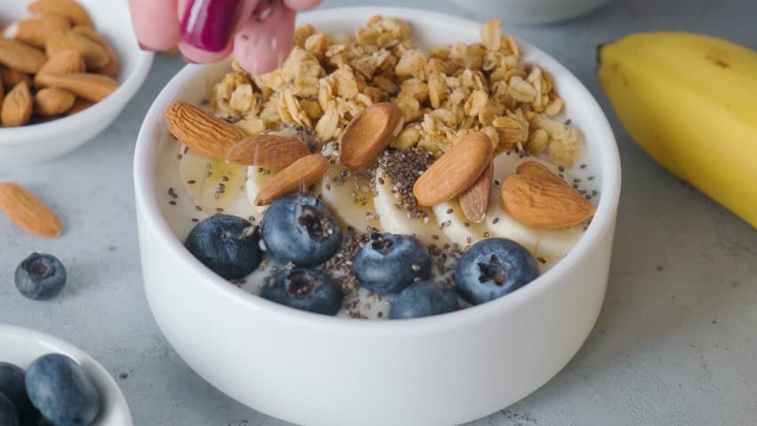 Woman hand add chia seeds into a bowl of greek yogurt with oat granola, banana, blueberries and almonds on gray background | Shutterstock HD Video #1103398493