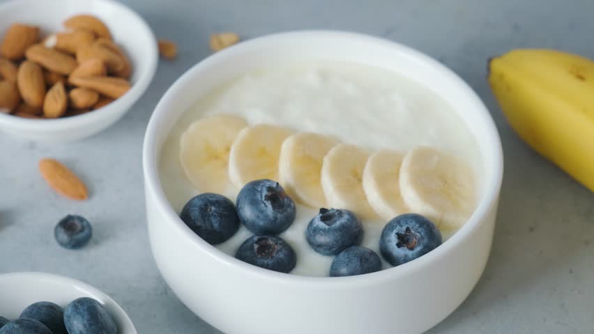 Woman hand add oat granola into a bowl of greek yogurt with banana, blueberries on gray background for cooking healthy breakfast Royalty-Free Stock Footage #1103398495