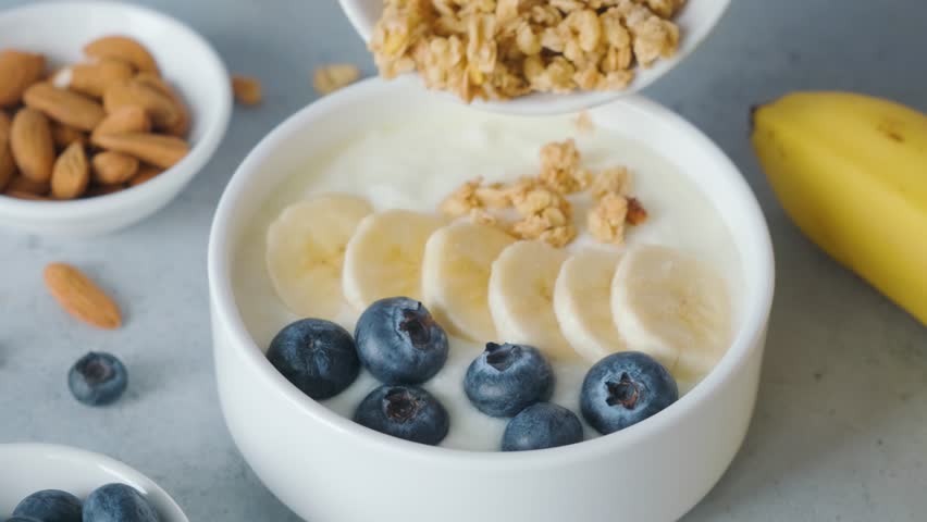 Woman hand add oat granola into a bowl of greek yogurt with banana, blueberries on gray background for cooking healthy breakfast | Shutterstock HD Video #1103398495