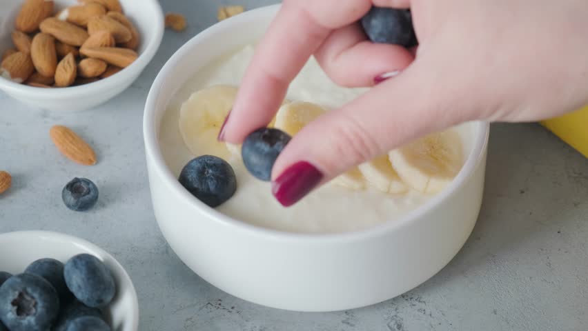 Female hand blueberries into a bowl of greek yogurt with banana on gray background for cooking healthy breakfast | Shutterstock HD Video #1103398497