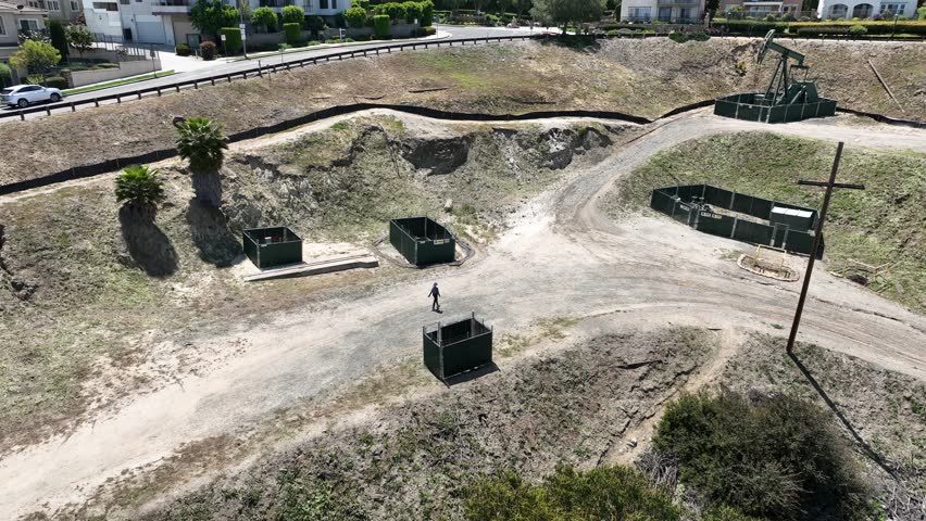 4K drone footage of a woman walking near decommissioned pumpjack-style oil wells while on a hiking path in Signal Hill, California. 