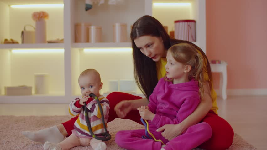 Portrait of caring charming mother with adorable infant baby and lovely little daughter sitting on floor, enjoying leisure and bonding while playing with colorful toys in domestic room.