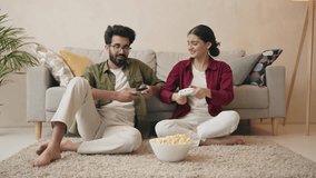 Indian young couple sitting on couch in living room enjoy playing video games spending time together. Happy couple play video games at home both are smiling, laughing and enjoying moment on weekend