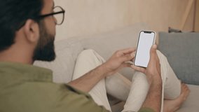Young male tech user relaxing on sofa holding smartphone mock up blank white screen. Man customer using cell phone for video call. Over shoulder view