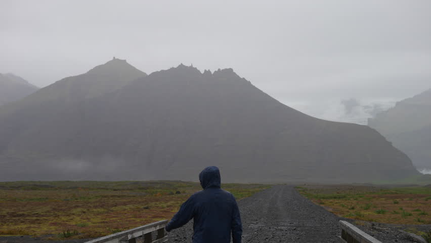 Back view shot of a man wearing a raincoat pointing his fingers while standing on a bridge in the middle of a road near a mountain in Iceland