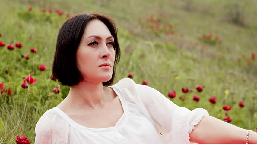 Portrait A pretty woman in a white blouse and a scarlet skirt sits in the grass among red wild peonies. Festival and holiday event