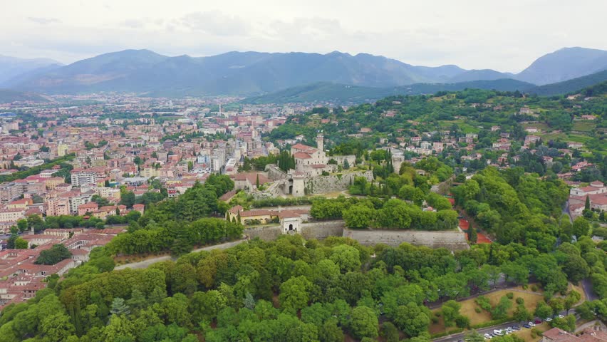 Inscription on video. Brescia, Italy. Medieval Castello di Brescia fortress on a hill with burning text overlay, overlooking the historic city under dramatic cloudy skies. Aerial view