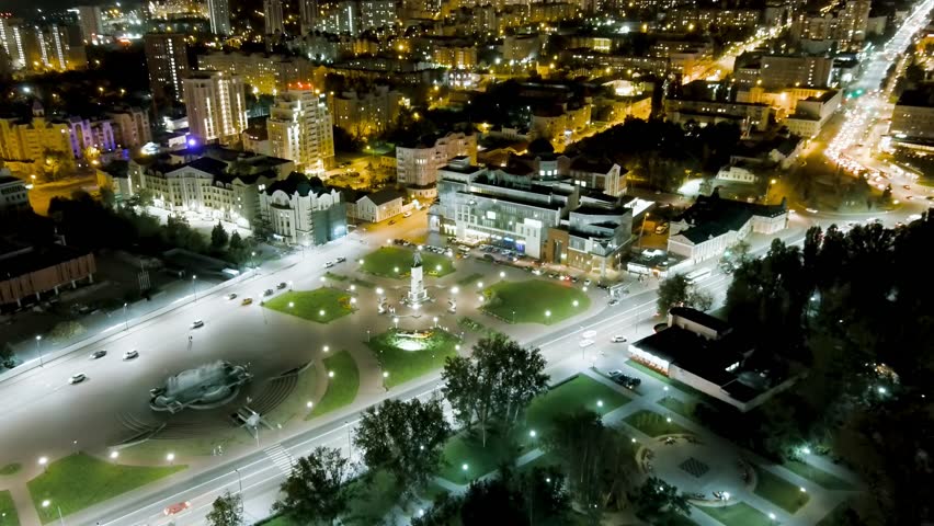Lipetsk, Russia. Peter the Great Square is seen from above in a sweeping aerial view after sunset, with the city center illuminated by thousands of golden streetlights and building lights. The square features a prominent monument at its center, surrounded by neatly arranged green lawns and pathways, with wide avenues carrying streams of vehicle traffic on either side. The surrounding architecture blends historic and modern structures, their rooftops and facades glowing against the darkening sky. The scene captures the vibrant energy of a regional Russian city at night, where urban thoroughfares converge on a central plaza dedicated to the emperor who founded the settlement. The monument was unveiled in 1996 as part of the 300th anniversary celebrations of the Russian Navy, which Peter the Great established. The camera gradually ascends and tilts, revealing an expanding panorama of the illuminated street grid stretching toward the horizon.