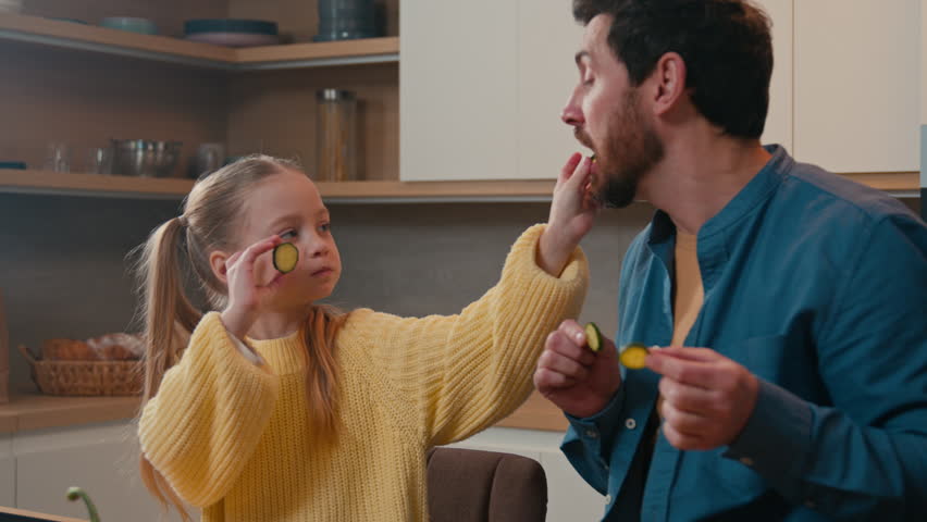 Caucasian father with little daughter at home kitchen cooking eating together child girl kid feed dad with cucumber slice daddy joking pretend feeding baby deceive cheat fooling around happy family