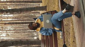 Female working on laptop in city park wearing medical face mask. Vertical video. Woman freelancer in casual clothes sitting on bench in park and using notebook, typing messages and sending letters