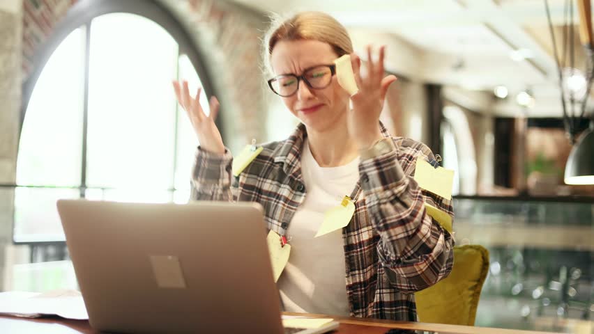 Nervous stressed young blond woman freelancer with stickers typing quickly on laptop computer and looking at paper documents asking to hurry up scream and warning about deadline at coworking workplace