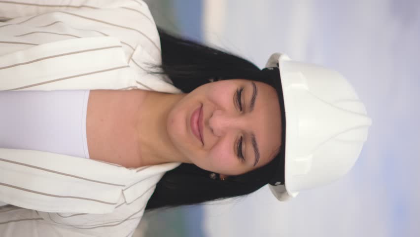 Young pretty girl in construction helmet shows OK sign with her fingers against background of modern line and mountain landscape. She smiles and looks at camera. advertising. vertical video