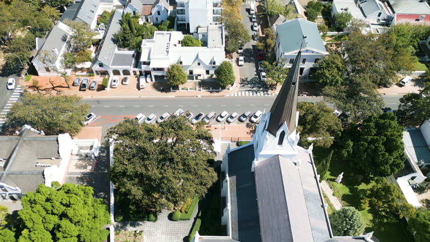 aerial downwards tracking shot of church spire and church building, NG Kerk, Stellenbosch with streets, people on sidewalks, cars, buildings, trees, sunny