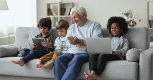 Positive multiethnic kid boys and senior elder granddad man resting on couch with gadgets together, using digital electronic devices for online communication, chatting, studying, playing on Internet