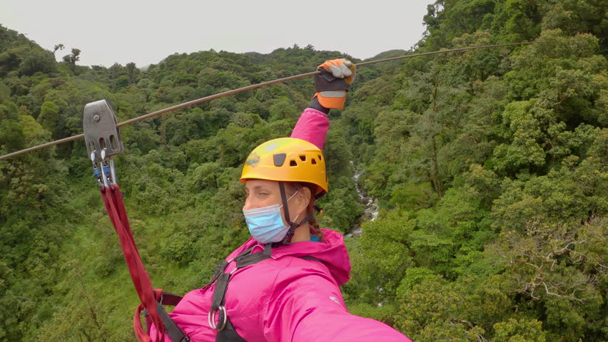 Young lady with face mask on zipline above lush jungle trees and wild river. While holidays in mountain town of Boquete in Chiriqui Province, she has an adrenaline experience in pristine rainforest.
