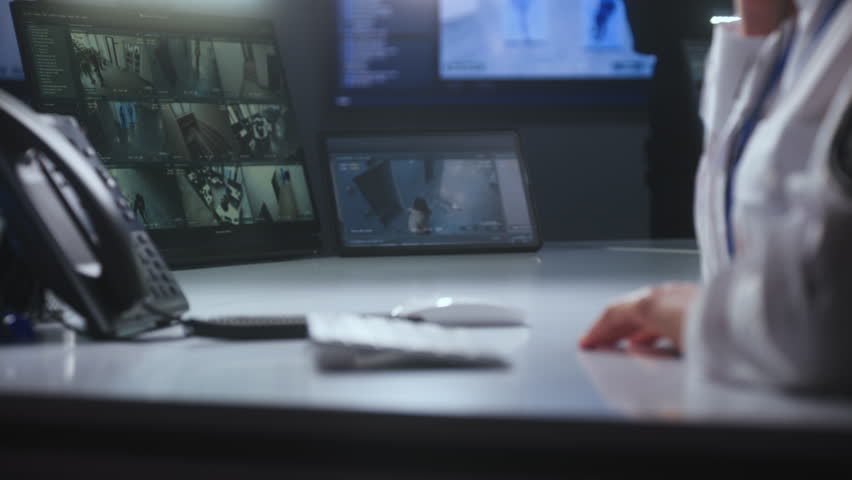 Female security officer works in police monitoring center. Woman types on keyboard, uses landline phone and radio set. Surveillance cameras view displayed on computer and tablet screens. Close up.