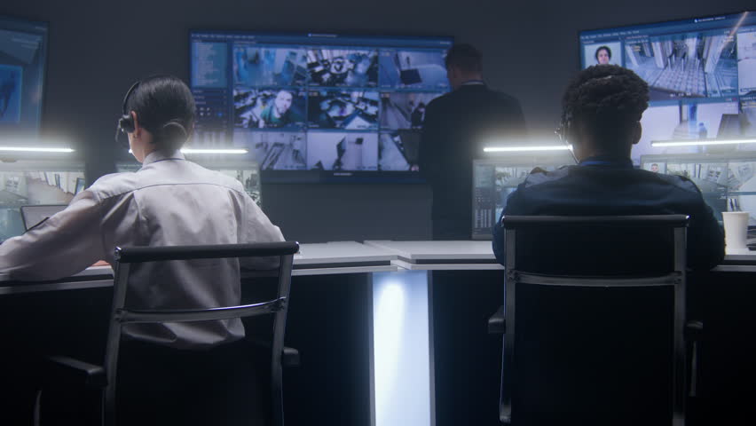 Multi ethnic security employees monitor CCTV cameras in police surveillance center. Multiple big screens on the wall and PC monitors showing security cameras footage. Tracking system. Social safety. Royalty-Free Stock Footage #1103472115