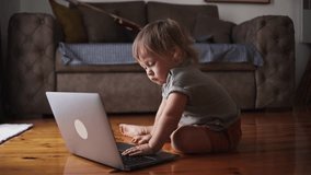 A one-year-old child is sitting in front of a laptop. Funny little kid is working on a laptop at home. The concept of learning, parenting, childhood, working remotely, programming