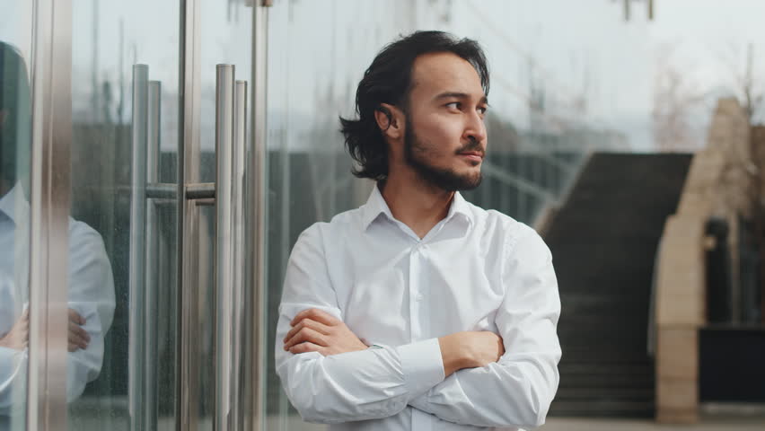 Young successful asian businessman in shirt, near the business center, folds arms and smiles, front view. Manager is standing outdoors with arms crossed over chest