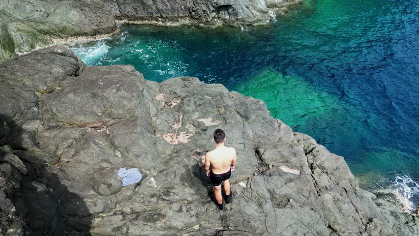 Aerial View of Shirtless Male Tourist Jumping off rocky cliffside into clear, turquoise ocean waters in tropical island of Catanduanes.