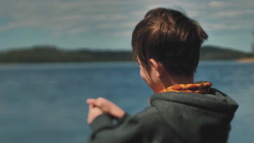 A little boy stands next to the lake and looks and waves. Active and smiling child resting alone Soft selective focus.