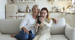 Happy pretty mature grandmother and teenage girl taking selfie on mobile phone together, using application for funny media effects, smiling, laughing, enjoying Internet communication at home