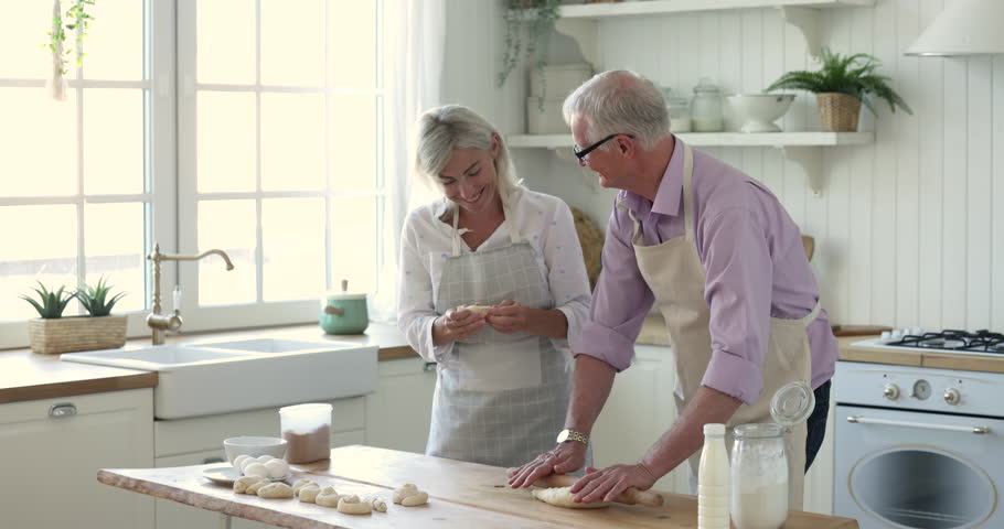 Happy elderly retired married couple baking in kitchen together, rolling, shaping dough, having fun, preparing pastry, bakery food, dessert, buns for breakfast, talking, smiling, laughing Royalty-Free Stock Footage #1103492009
