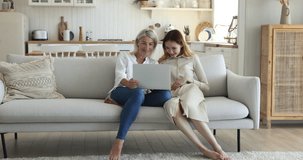 Positive teenage kid girl and mature grandmother using laptop at home together, resting close on home sofa, watching content, making video call, looking at screen, speaking, smiling, laughing