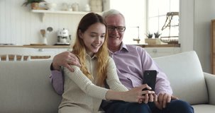 Cheerful teen grandkid girl and happy positive grandpa taking selfie picture on mobile phone at home, sitting on couch together, laughing, smiling, talking, grimacing on cellphone screen