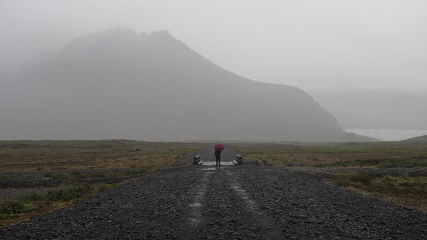 A man clutching an umbrella stands on a small bridge in the middle of a country road, with a view of a large mountain in the distance