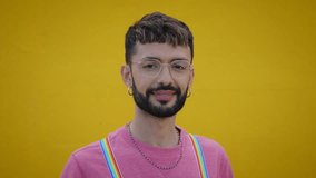 Close up smiling cheerful young Caucasian man with rainbow lgbt suspenders. Gay people posing for video yellow background. Cheerful gay bearded person happy studio isolated. Millennials generation.