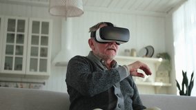 Excited mature senior man wearing using virtual reality metaverse VR glasses headset at home. Grandfather touching air during VR experience on virtual reality helmet. Simulation hi-tech videogame