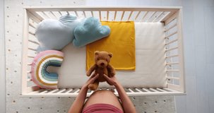 Pregnant woman preparing nursery showing belly baby bump by crib putting teddy bear in crip. Pregnancy concept and home nusery planning. Top view video in slow motion