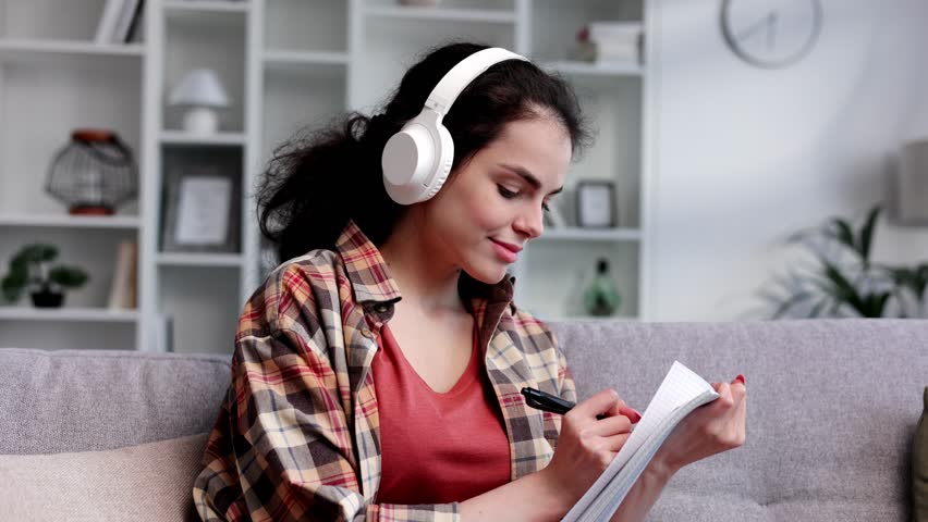 Curly-haired gir studentl sits on the sofa, wears headphones, writes with a pen in a notebook and checks notifications on the phone. A beautiful woman studies and switches the music that will play