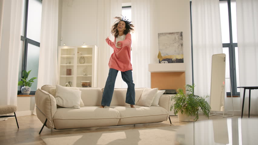 Excited happy young woman jumping happily on modern sofa. Afro girl homeowner having fun on move in day, celebrating mortgage approval, relocation to upgraded apartment. Bank mortgage concept