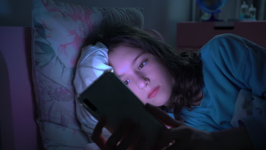 Teen girl lying in bed at night using smartphone, scrolling social media and texting. Not healthy sleep. Insomnia and the harm of the smartphone before going to bed. Dependence on social networks