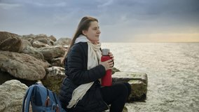 Woman with long hair enjoying a hot cup of tea while sitting in the beautiful sea coastline on cold windy day. Great for use in vacation and travel videos