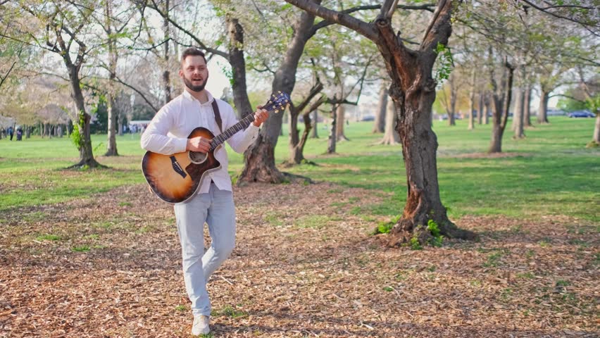 Adult man and musician, singer, guitarist, and artist is walking through a park singing a song and strumming his guitar