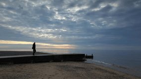 Slow motion wide shot of man walking to end of jetty at dusk on Fleetwood Beach Lancashire UK