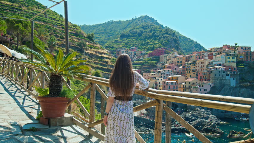Female tourist walking in Riomaggiore harbor looking at quaint village overhanging cliffs. Cinque Terre with a small harbor beach and a wharf framed by colorful tower houses in Italy. Royalty-Free Stock Footage #1103542685