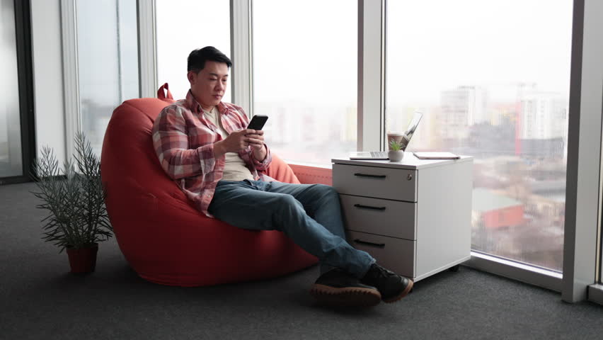 Serious asian male worker taking little break and surfing internet on smartphone. Young man in casual clothes having rest on soft pouffe chair near panoramic windows in modern comfortable office. Royalty-Free Stock Footage #1103549137