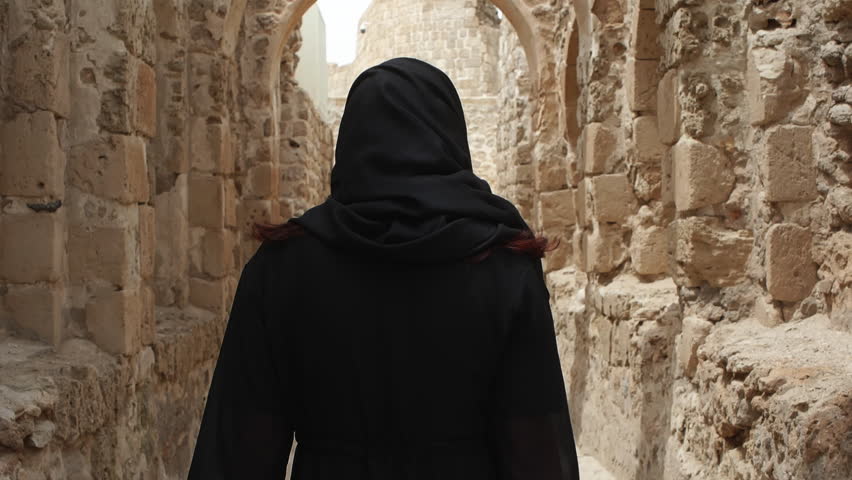 Woman in arabian black hijab headscarf walks in Bahrain Fort under arch, rear view. Archaeological site from stones sand, conservation of traditions and culture. Tourism, travel, tour, journey. Royalty-Free Stock Footage #1103550723