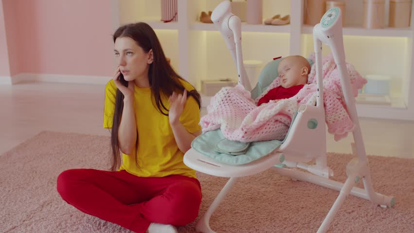 Attractive positive mother sitting on floor , taking a rest of daily routine , chatting on smartphone with friend while peaceful adorable infant daughter sleeping in electric cradle baby swing indoors