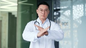 Asian doctor in white coat showing crossed arms, X sign, shaking head no while standing in modern hospital clinic. Medical worker physician demonstrates a gesture of prohibition, denial, disagreement - Powered by Shutterstock - Get 15% off with code: PIKWIZARD15