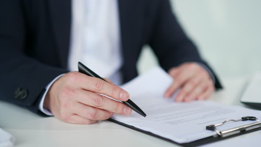 Close up of a man's hand signing documents at a desk at a workplace in a modern office. The boss owner in a formal suit looks through the folder with documentation and signs the contract with a pen Royalty-Free Stock Footage #1103553881
